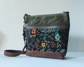 Puede incluir: Un bolso bandolera con un panel de tela negra con estampado floral colorido, una sección superior de lona verde oliva y una base de cuero marrón. El bolso incluye un bolsillo frontal con cremallera, cierre superior de cremallera y una correa ajustable.