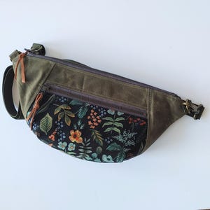 Könnte beinhalten: Eine grüne und schwarze Bauchtasche mit Blumendruck. Die Tasche hat eine Reißverschlusstasche und einen braunen Lederriemen.