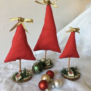 Könnte beinhalten: Drei rote Stoff-Weihnachtsbäume mit goldenen Punkten, jeder mit einer goldenen Schleife und Glocke. Die Bäume stehen auf Holzsockeln mit Grün und kleinen Geschenkboxen. Mehrere Weihnachtsbaumkugeln in Rot, Gold und Grün sind verstreut.