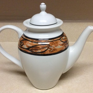 Może przedstawiać: Biały ceramiczny czajnik z brązowym i czarnym wzorem liści wokół środka. Czajnik ma pokrywkę z gałką na górze.