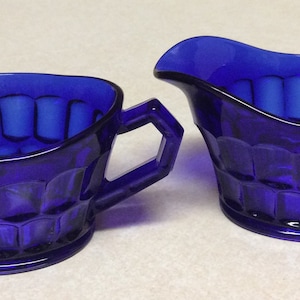 May include: A cobalt blue creamer and sugar bowl set with a textured design. The creamer has a handle on the right side and the sugar bowl has a handle on the left side.
