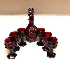 Puede incluir: Un juego de decantador y copas de vino de vidrio rojo rubí. El decantador y las copas presentan un diseño texturizado. Seis copas están dispuestas alrededor del decantador.