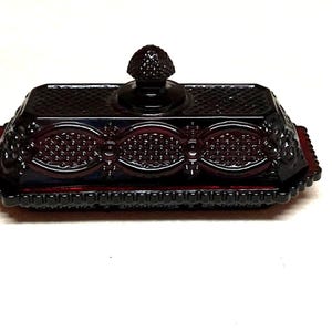 Puede incluir: Una mantequera de cristal rojo rubí con tapa y pomo decorativo. El plato tiene forma rectangular con patrones en relieve ornamentados y borde festoneado. La tapa presenta un diseño texturizado.