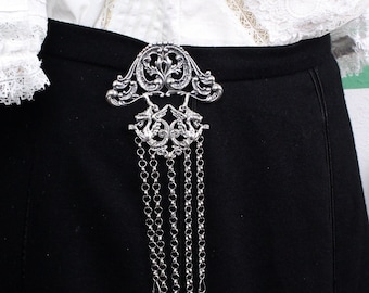 May include: A silver-toned belt buckle with an ornate design and dangling chains is attached to a black skirt. The buckle is the focal point, set against the dark fabric. A white blouse with lace trim is visible.