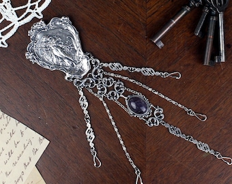 May include: A silver-toned, ornate hand-shaped jewelry piece with an oval, dark purple stone. The piece has intricate chain details and decorative elements. A collection of antique keys rests nearby on a dark wooden surface.