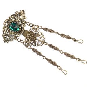 Puede incluir: Un colgante adornado en tonos dorados con una piedra preciosa verde en el centro y tres cadenas colgantes con extremos decorativos.