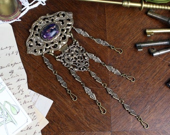 May include: An antique-style brooch with an ornate, filigree design in a bronze tone. The brooch features a central oval stone with purple and gold flecks. Dangling chains with decorative elements and hook closures complete the piece.