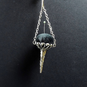 Può includere: Un cuscino per spilli in argento con un top in tessuto nero appeso a una catena d'argento. Il cuscino per spilli ha la forma di un cono con una parte superiore decorativa e ornata.