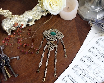 May include: A gold-toned pendant with a green gemstone and intricate filigree details. The pendant hangs from a chain with multiple loops.