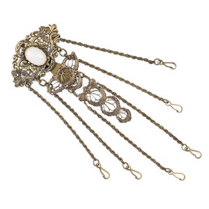 Puede incluir: Un accesorio para el cabello de estilo antiguo con una piedra ovalada opalescente engastada en una base de metal filigranado ornamentado. La pieza presenta cadenas colgantes con cierres de gancho, probablemente de latón.