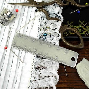 Op de afbeelding: Een verzameling naai-attributen, waaronder een metalen liniaal, vingerhoed, spelden en schaar. De liniaal meet tot 15 cm. Witte stof met zwarte strepen en kanten afwerking, wat duidt op naaien.