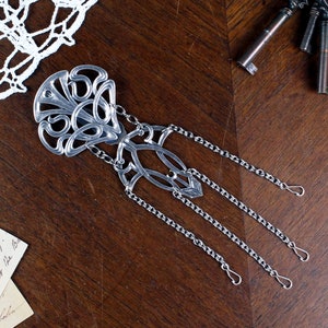 Può includere: Un ciondolo in metallo argentato con un design decorativo e intricato. Il ciondolo è attaccato a quattro catene argentate con un gancio alla fine di ogni catena.