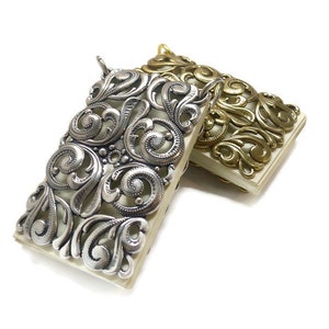 Könnte beinhalten: Zwei verzierte Metall-Buch-Anhänger, einer silberfarben und einer golden, mit filigranen Designs. Die Anhänger sind rechteckig mit einem dekorativen Rand.
