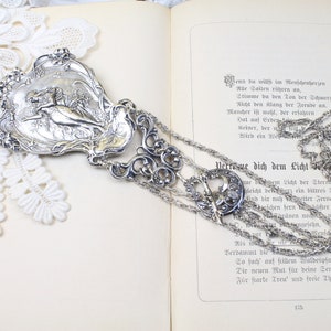 Può includere: Una collana argentata ornata con un pendente dettagliato raffigurante una scena a rilievo classica. La collana ha più fili di catena delicata ed è esposta su un libro aperto con testo tedesco.