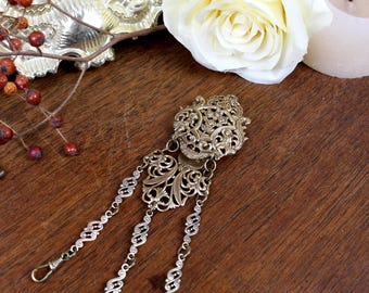 May include: A gold-toned metal brooch featuring an intricate filigree design with a central oval plate and two smaller decorative sections. Three delicate, patterned metal chains hang from the base, each ending in a small, simple hook clasp.