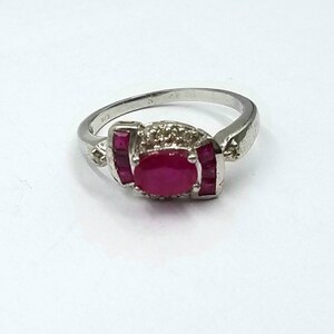 Könnte beinhalten: Ein silberner Ring mit einem großen ovalen Rubin in der Mitte, flankiert von kleineren rechteckigen Rubinen. Der Ring hat eine detaillierte Fassung mit kleinen Steinen, die ein elegantes Design schaffen. Der Ring ist vor einem weißen Hintergrund platziert.