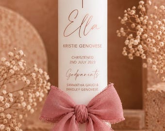 May include: A white pillar candle featuring a rose gold cross and the name Ella, followed by Kristie Genovese, the date 2nd July 2023, and the names of godparents Samantha Gauci and Bradley Genovese, tied with a dusty rose fabric bow.