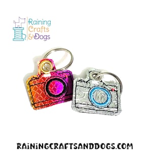 May include: Two camera-shaped keychains, one with a holographic iridescent finish and the other with a silver finish. Both keychains have a blue circle in the center of the camera lens.