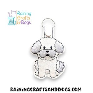 Könnte beinhalten: Weißer bestickter Schlüsselanhänger mit einem Cartoon-Pudel in Weiß mit grauen Ohren und schwarzer Nase. Der Schlüsselanhänger ist an einem silbernen Schlüsselring befestigt.