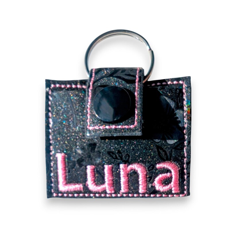 May include: A square black keychain featuring a subtle floral pattern with glittery accents. The keychain has a snap closure, pink embroidered stitching along the edges, and the name Luna embroidered in pink thread on the front surface.