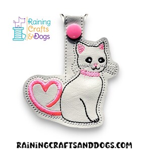 May include: A gray cat-shaped keychain with a pink heart tail and collar. The cat is embroidered with black thread and has a pink button at the top. The text "Raining Crafts & Dogs" is visible.