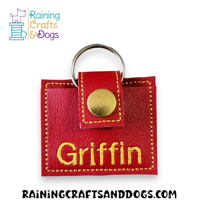 May include: A square red faux leather keychain featuring a gold-colored metal snap closure and a silver-colored metal keyring. The name Griffin is embroidered in yellow thread on the front. The logo Raining Crafts & Dogs is visible.
