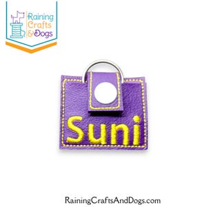 May include: A square purple faux leather keychain featuring the name Suni embroidered in yellow thread. The item includes a silver metal split ring at the top and a small flap secured with a white plastic snap button, finished with yellow stitching.