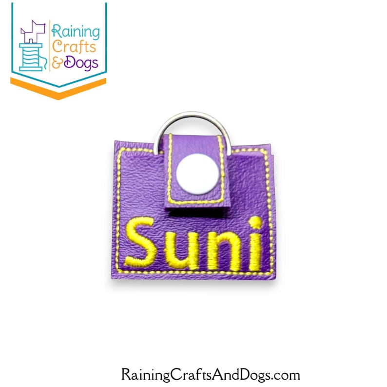 May include: A square purple faux leather keychain featuring the name Suni embroidered in yellow thread. The item includes a silver metal split ring at the top and a small flap secured with a white plastic snap button, finished with yellow stitching.