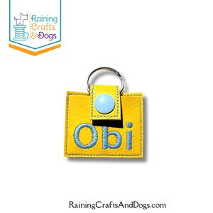 May include: A square yellow keychain made of a textured material featuring the name Obi embroidered in light blue thread. The keychain includes a light blue snap button closure and a silver metal split ring attached to the top center.