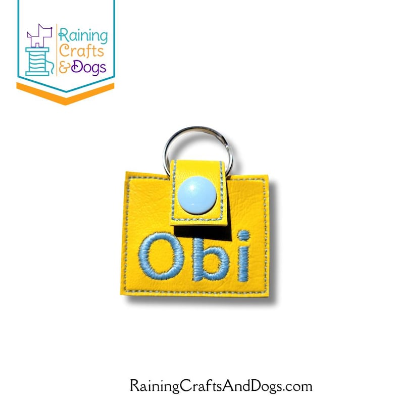 May include: A square yellow keychain made of a textured material featuring the name Obi embroidered in light blue thread. The keychain includes a light blue snap button closure and a silver metal split ring attached to the top center.