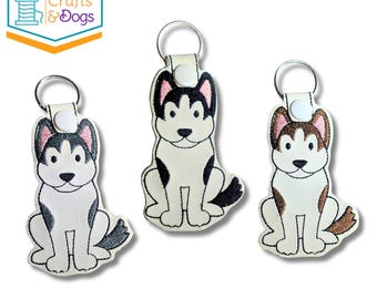 May include: Three white faux leather keychains featuring embroidered husky dog designs. Two keychains depict black and white huskies, while one features a brown and white husky. Each keychain includes a silver metal ring and a white snap button.