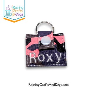May include: A square keychain featuring a pink, black, and dark gray camouflage pattern. The keychain has a white snap closure on a small tab, a silver metal ring, and the name Roxy embroidered in white thread across the front surface.