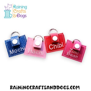 May include: Four rectangular faux leather keychains in blue, light pink, red, and dark pink. Each features a white snap button, a silver metal keyring, and white embroidered names: Mochi, Hachi, Chibi, and Zuki Bean. The logo Raining Crafts & Dogs is visible.