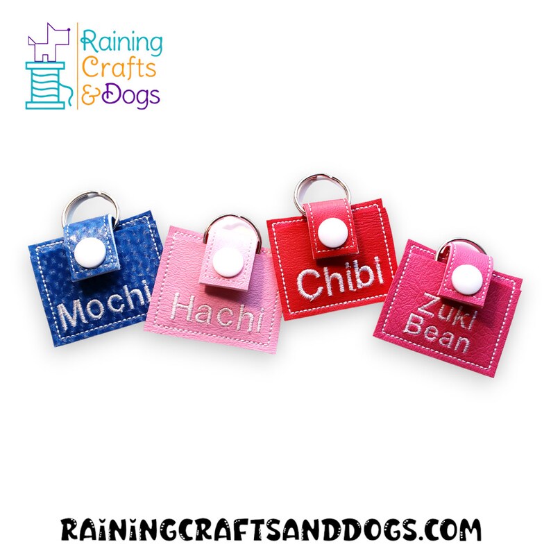 May include: Four rectangular faux leather keychains in blue, light pink, red, and dark pink. Each features a white snap button, a silver metal keyring, and white embroidered names: Mochi, Hachi, Chibi, and Zuki Bean. The logo Raining Crafts & Dogs is visible.