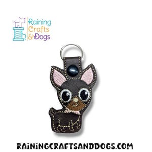 Puede incluir: Un llavero de piel sintética marrón oscuro con forma de perro chihuahua con detalles bordados, incluyendo grandes ojos negros, hocico color canela, orejas internas rosas y un pequeño lazo rosa. Un anillo de metal plateado está unido a la parte superior.