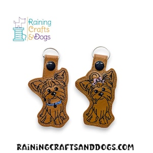 Puede incluir: Dos llaveros de cuero marrón con diseños de Yorkshire Terrier bordados. Un perro lleva un collar azul y el otro un lazo rosa.