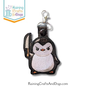 May include: A penguin-shaped keyring in black and white, with an angry face and a knife. It has a silver clasp and a black leather strap. The text "Raining Crafts & Dogs" and "RainingCraftsAndDogs.com" are also visible.