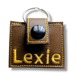 May include: A square brown faux leather keychain featuring a silver metal ring at the top. The front is embroidered with the name Lexie in yellow thread, accented by yellow stitching along the edges and a black snap button closure on the tab.