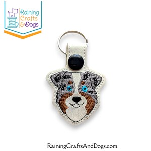 May include: A white faux leather keychain featuring an embroidered Australian Shepherd dog face with blue eyes, grey and brown fur details, and a black nose. The keychain includes a silver metal ring and a black snap button closure at the top.