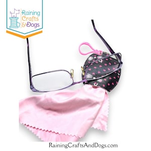 Puede incluir: Gafas con montura morada con un paño de limpieza rosa y un estuche negro para gafas con un patrón de corazones rosas. La funda tiene un clip rosa. El logotipo "Raining Crafts & Dogs" está en la esquina superior izquierda.