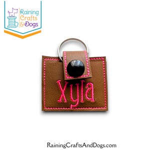 May include: A rectangular brown faux leather keychain featuring bright pink embroidered stitching around the edges and the name Xyla embroidered in pink. A black snap button secures the flap, and a silver metal ring is attached to the top.