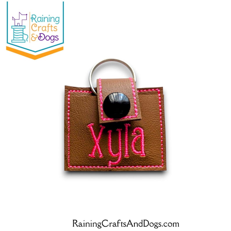 May include: A rectangular brown faux leather keychain featuring bright pink embroidered stitching around the edges and the name Xyla embroidered in pink. A black snap button secures the flap, and a silver metal ring is attached to the top.