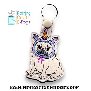 May include: White pug wearing a blue unicorn costume with a rainbow horn and pink bow. The pug is sitting and looking to the right. The costume has white fur trim. The pug is attached to a white key ring with a silver key ring loop.