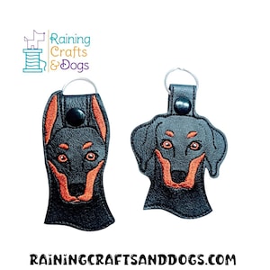 Puede incluir: Dos llaveros de cuero negro con cabezas de perro Doberman Pinscher bordadas. Los ojos del perro son de color naranja y la nariz y la boca son rojas.