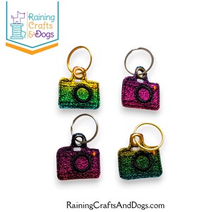 May include: Four camera-shaped keychains featuring embroidered details and metallic split rings. Each keychain displays a gradient of colors including green, yellow, pink, and purple with black stitching around the lens and body edges.