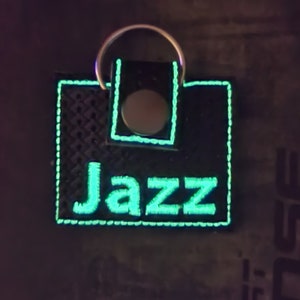 May include: A square black keychain with a textured surface featuring the word Jazz embroidered in glowing light green thread. The keychain has a matching light green stitched border and a silver metal snap button closure with a circular ring attached.