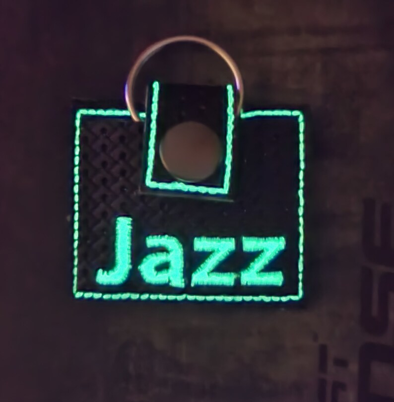 May include: A square black keychain with a textured surface featuring the word Jazz embroidered in glowing light green thread. The keychain has a matching light green stitched border and a silver metal snap button closure with a circular ring attached.