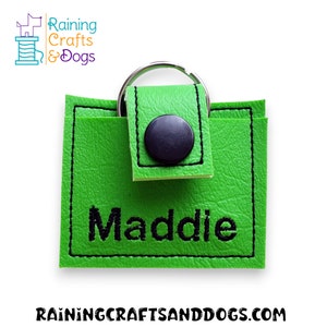 May include: A square, bright green keychain pouch featuring a black snap closure and black stitching along the edges. The name Maddie is embroidered in black thread on the front. A silver metal keyring is attached to the top of the pouch.
