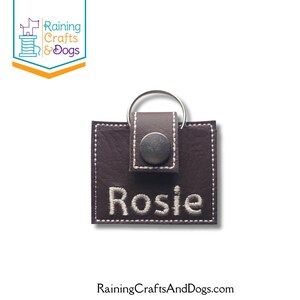 May include: A square dark brown faux leather keychain featuring white contrast stitching and a silver metal snap button. The name Rosie is embroidered in white thread on the front. A silver metal split ring is attached to the top tab.