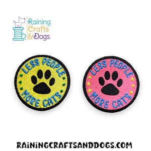 May include: Two circular embroidered patches with black borders. One patch has a yellow background, the other a pink background. Both feature a black cat paw print in the center and the blue embroidered text "LESS PEOPLE MORE CATS" around the edge.